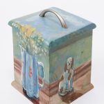 A small biscuit box painted on all sides and the lid with flowers in a jug and a dog ornament.